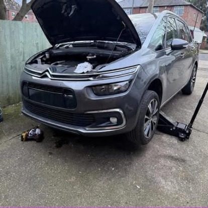 A grey Citroen C4 Grand Picasso on a residential driveway with the bonnet open, a trolley jack under the front and an oil drain pan on the floor.