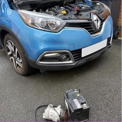A blue Renault Captur with the bonnet up, its removed alternator and battery standing together on the ground in front of the bumper.