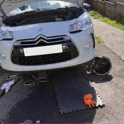 A white Citroen DS3 on a driveway with the bonnet up and its gearbox lying on rubber matting beside the front wheel, next to a trolley jack and a pair of orange gloves.