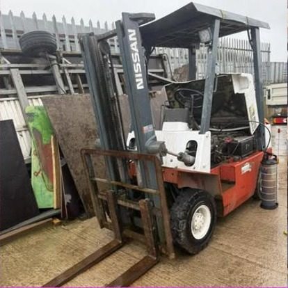 A red and white Nissan forklift truck standing in a yard with its engine cover lifted, palisade fencing and stacked steel behind it.