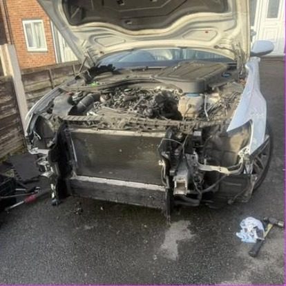 A white car on a driveway with its entire front end removed down to the radiator pack and crash bar, bumper and headlights off, tools laid out on the ground.