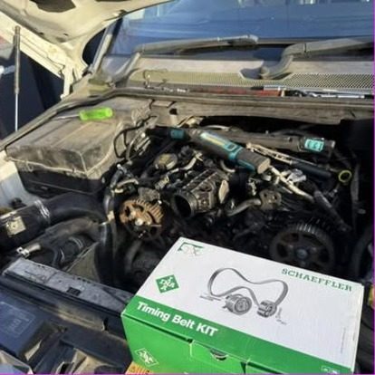 An engine bay opened up with the timing cover removed and the belt and pulleys exposed, a boxed Schaeffler timing belt kit standing on the slam panel.